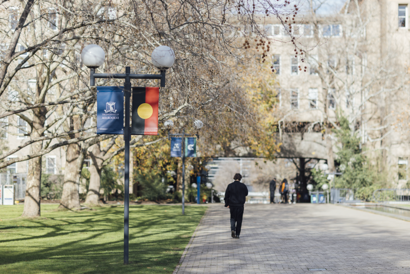 Student walking down a path at UoM Campus Parkville with Aboriginal and Torres Strait Island Flags to the side
