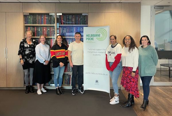 2023 cohort standing with Associate Professor Michelle KennedyExecutive Manager, Research and Knowledge Translation, Lowitja Insitute, in front of a Melbourne Poche banner 